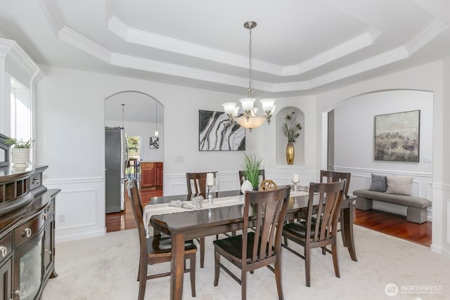 dining space with a wainscoted wall, light wood-type flooring, arched walkways, a notable chandelier, and a raised ceiling