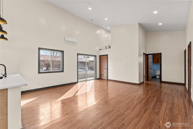 unfurnished living room with wood finished floors, baseboards, recessed lighting, an AC wall unit, and a towering ceiling