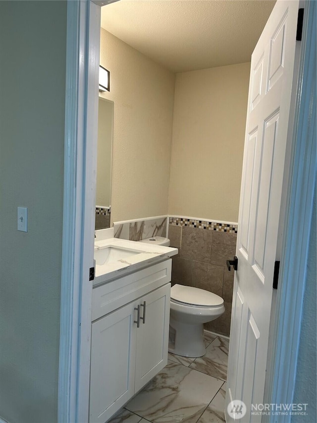 half bath featuring vanity, a textured ceiling, wainscoting, toilet, and marble finish floor