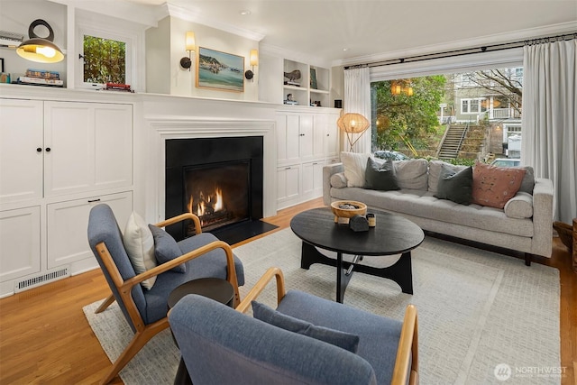 living room with built in features, visible vents, a fireplace with flush hearth, light wood-style floors, and crown molding