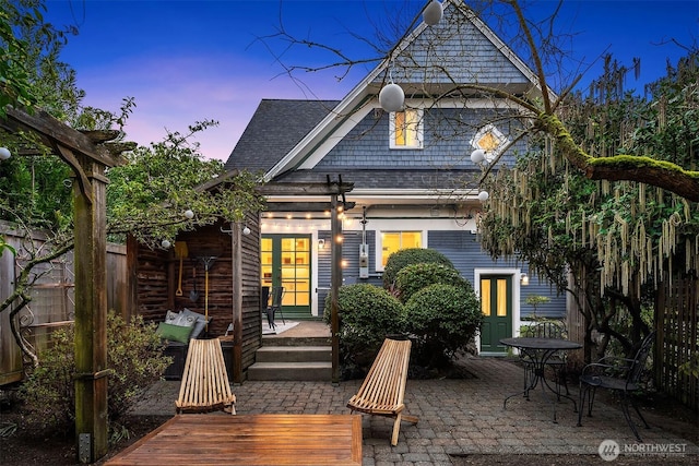 back of house at dusk featuring a patio area, french doors, fence, and a shingled roof