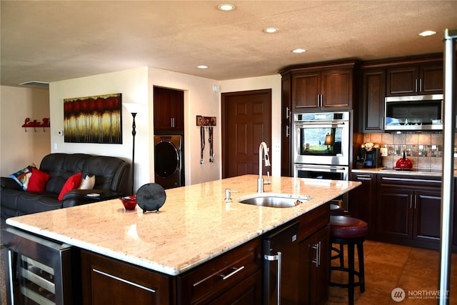 kitchen with a kitchen island with sink, a sink, wine cooler, appliances with stainless steel finishes, and tasteful backsplash