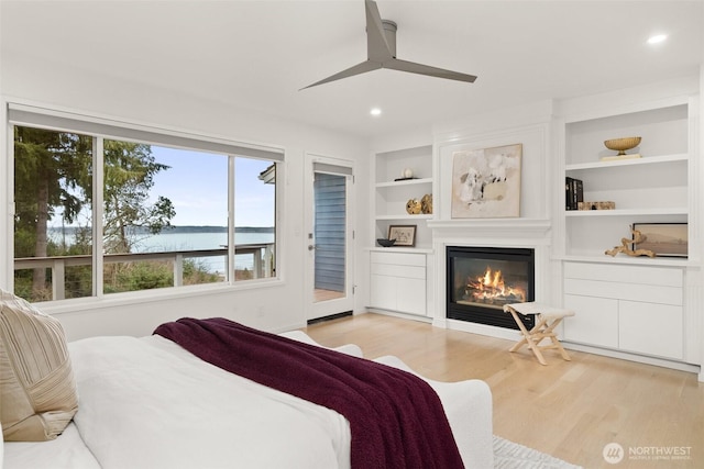 bedroom featuring ceiling fan, a water view, light wood-style flooring, a glass covered fireplace, and access to outside