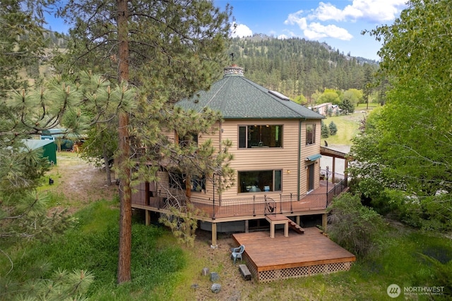 back of house featuring a view of trees, a deck, and roof with shingles