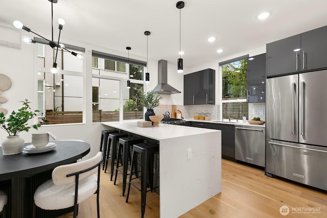 kitchen with wall chimney range hood, stainless steel appliances, light countertops, a kitchen breakfast bar, and light wood-type flooring