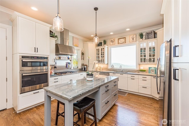 kitchen featuring a sink, wall chimney range hood, a kitchen island, appliances with stainless steel finishes, and light wood finished floors