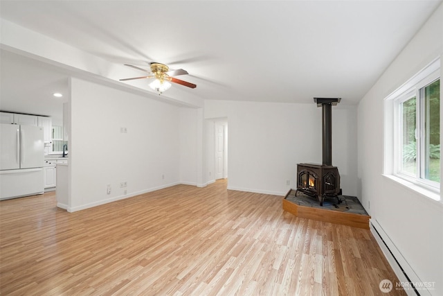 unfurnished living room featuring baseboards, a wood stove, light wood-style flooring, ceiling fan, and baseboard heating