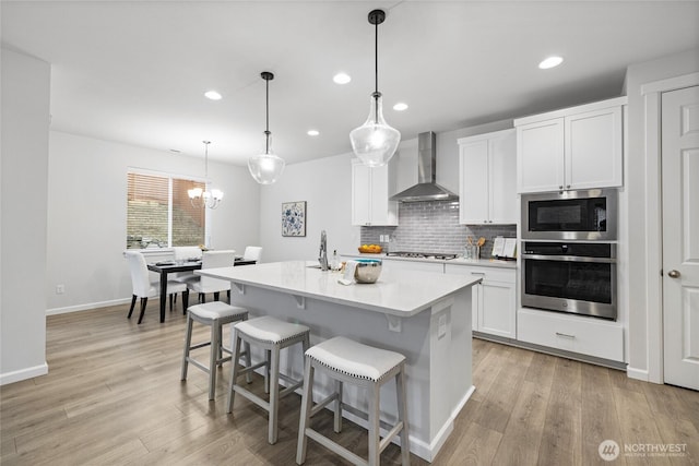 kitchen with tasteful backsplash, an island with sink, light wood-style flooring, appliances with stainless steel finishes, and wall chimney exhaust hood