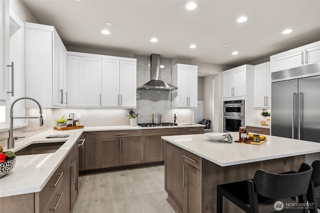 kitchen with light wood finished floors, wall chimney range hood, a kitchen breakfast bar, appliances with stainless steel finishes, and a sink