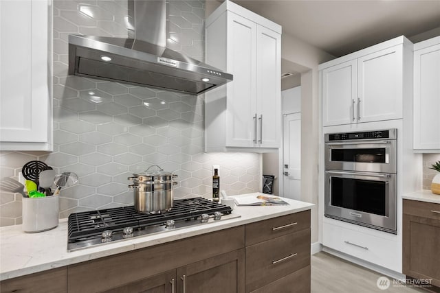 kitchen with light stone countertops, decorative backsplash, white cabinets, stainless steel appliances, and wall chimney exhaust hood