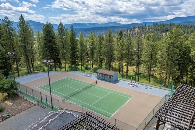 view of tennis court with a mountain view, an outdoor structure, a wooded view, and fence