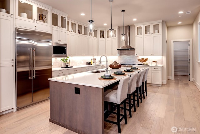 kitchen featuring light wood-style flooring, a kitchen island with sink, a sink, wall chimney range hood, and built in appliances