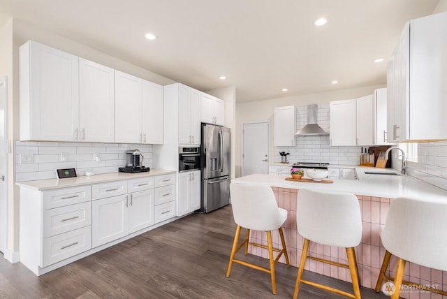 kitchen with a peninsula, a sink, stainless steel appliances, dark wood-type flooring, and wall chimney range hood