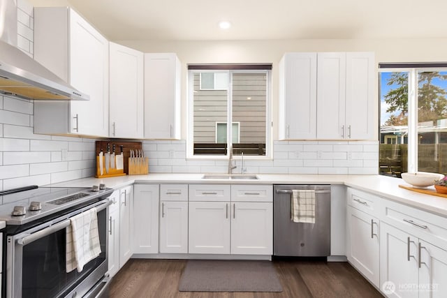 kitchen featuring a sink, appliances with stainless steel finishes, white cabinets, wall chimney range hood, and dark wood-style flooring