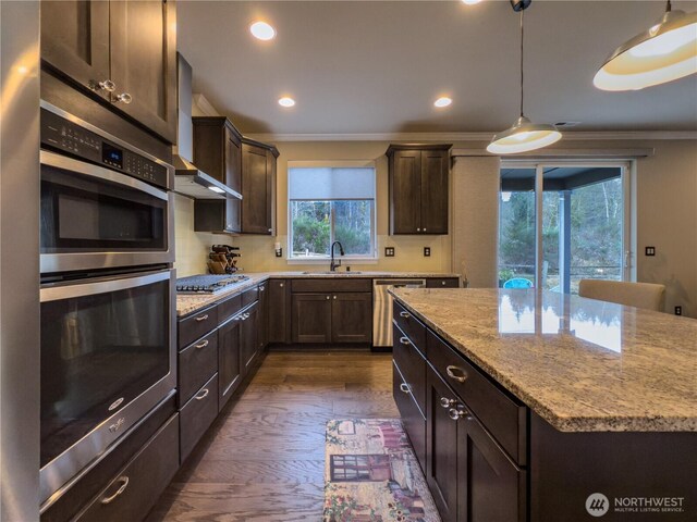 kitchen featuring a sink, light stone counters, dark brown cabinetry, appliances with stainless steel finishes, and wall chimney exhaust hood