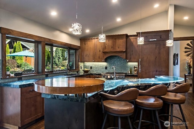 kitchen featuring lofted ceiling, a kitchen island with sink, dark wood-style flooring, hanging light fixtures, and backsplash