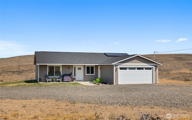 ranch-style home featuring a shingled roof, gravel driveway, roof mounted solar panels, covered porch, and a garage