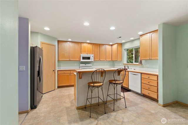 kitchen with light brown cabinetry, light countertops, recessed lighting, a kitchen breakfast bar, and white appliances