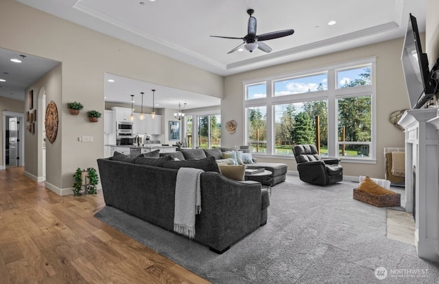 living room featuring light wood finished floors, baseboards, a fireplace with flush hearth, ceiling fan with notable chandelier, and a raised ceiling