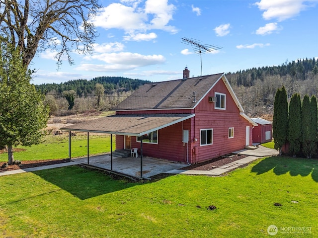 back of property with a forest view, roof with shingles, a chimney, a yard, and a patio area