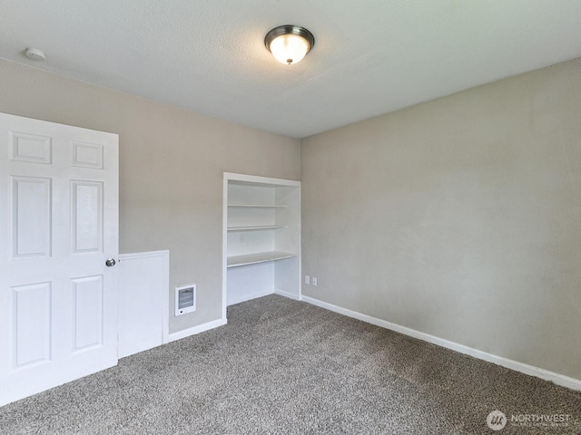 empty room featuring built in shelves, heating unit, a textured ceiling, dark colored carpet, and baseboards