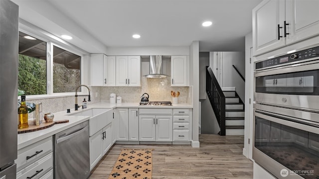 kitchen featuring light wood-style flooring, a sink, stainless steel appliances, white cabinets, and wall chimney range hood