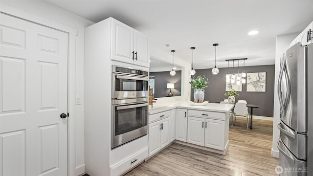 kitchen featuring appliances with stainless steel finishes, a peninsula, light wood-style floors, hanging light fixtures, and white cabinetry