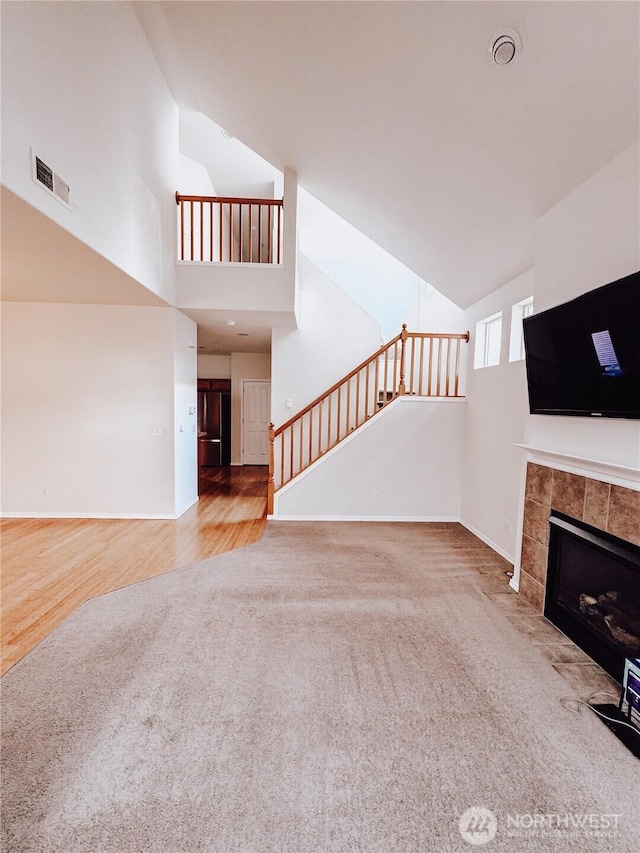 unfurnished living room with visible vents, stairs, carpet floors, a tile fireplace, and high vaulted ceiling