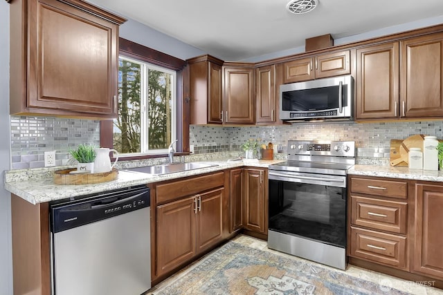 kitchen with tasteful backsplash, light stone counters, appliances with stainless steel finishes, brown cabinetry, and a sink