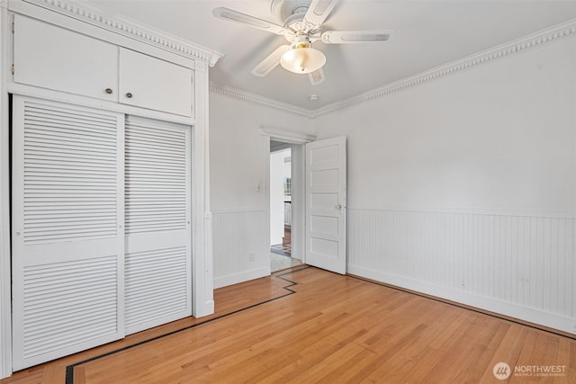 unfurnished bedroom with light wood-type flooring, ornamental molding, a ceiling fan, a closet, and wainscoting