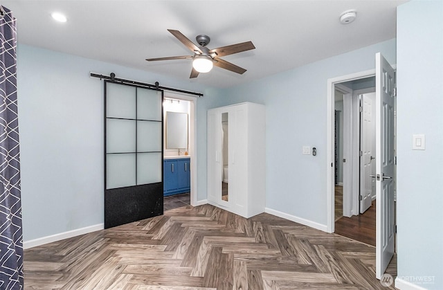 unfurnished bedroom featuring ceiling fan, a barn door, baseboards, and ensuite bath