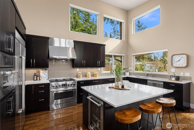 kitchen with a breakfast bar, wine cooler, appliances with stainless steel finishes, wall chimney range hood, and dark cabinets