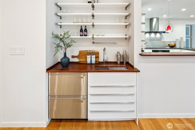 bar with a sink, wall chimney range hood, fridge, light wood finished floors, and wet bar