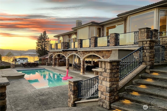 back of house at dusk with a patio, a balcony, an outdoor pool, and stone siding