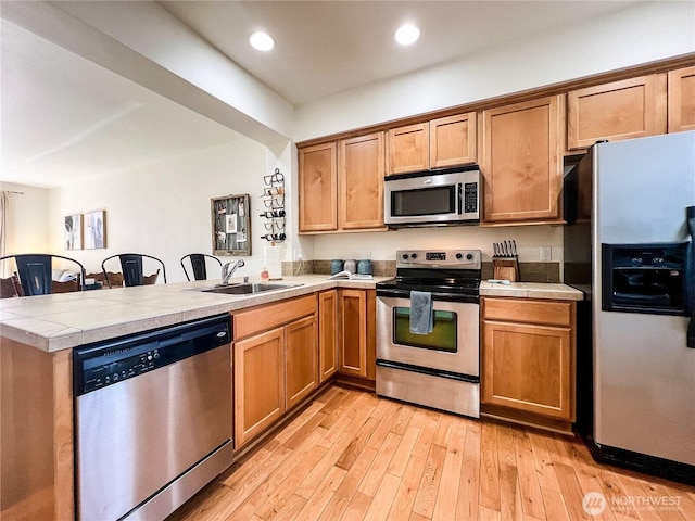 kitchen featuring tile counters, appliances with stainless steel finishes, a peninsula, light wood-style floors, and a sink