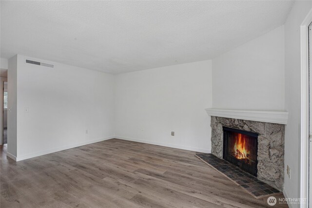 unfurnished living room with visible vents, baseboards, a stone fireplace, wood finished floors, and a textured ceiling