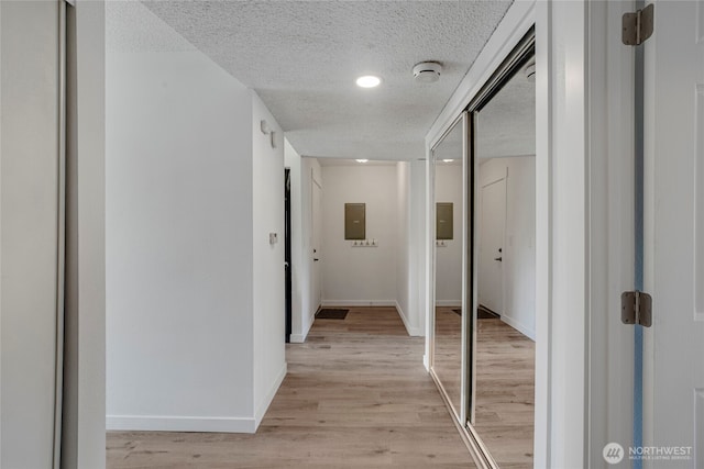 corridor featuring light wood finished floors, a textured ceiling, electric panel, and baseboards