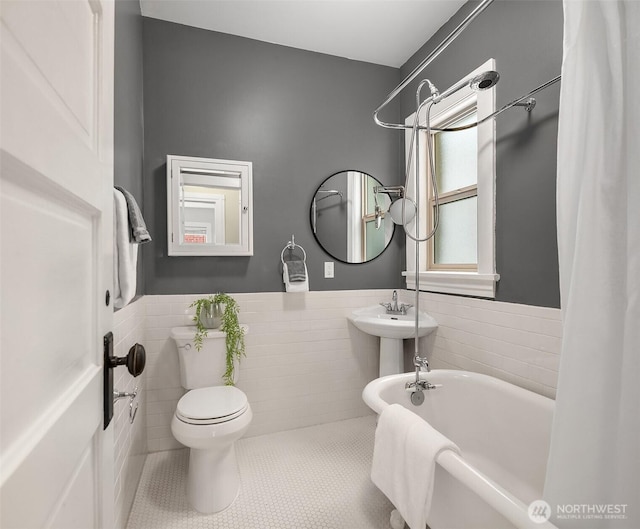 bathroom featuring a wainscoted wall, toilet, tile patterned floors, a soaking tub, and tile walls