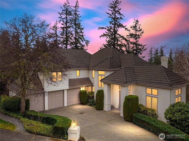 view of front facade featuring driveway, an attached garage, a chimney, stucco siding, and a tiled roof