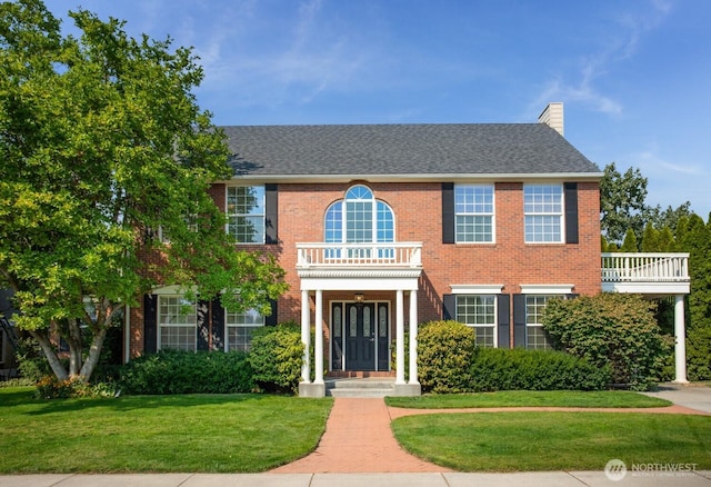 view of front of house with brick siding, a front lawn, roof with shingles, a chimney, and a balcony