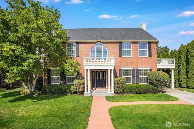 view of front of property with driveway, a balcony, a front yard, brick siding, and a chimney