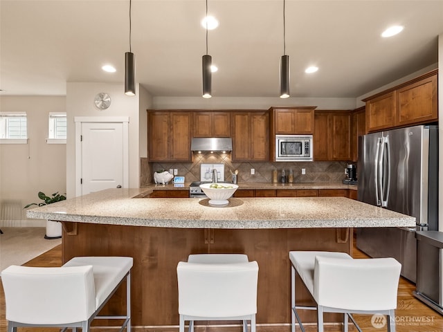 kitchen featuring decorative backsplash, under cabinet range hood, appliances with stainless steel finishes, and a breakfast bar