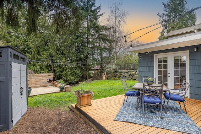 deck at dusk with outdoor dining space, fence, a yard, french doors, and an outdoor structure