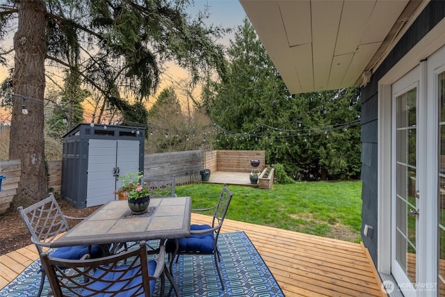 deck at dusk featuring a lawn, a fenced backyard, a storage shed, an outdoor structure, and outdoor dining area