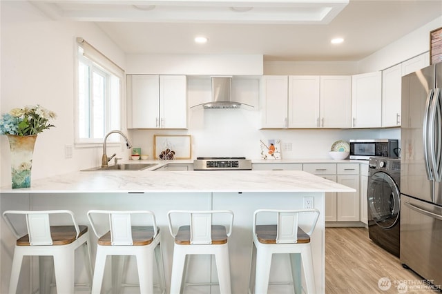 kitchen featuring a sink, washer / clothes dryer, appliances with stainless steel finishes, a peninsula, and wall chimney range hood