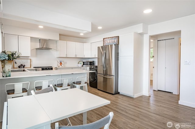 kitchen featuring light wood-type flooring, a peninsula, stainless steel appliances, washer / clothes dryer, and wall chimney exhaust hood