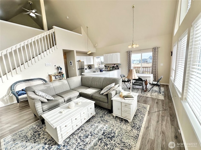 living area featuring high vaulted ceiling, ceiling fan with notable chandelier, stairway, light wood-style floors, and baseboards