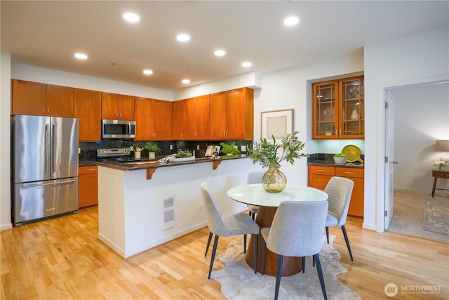 kitchen featuring a peninsula, light wood-style flooring, decorative backsplash, glass insert cabinets, and appliances with stainless steel finishes