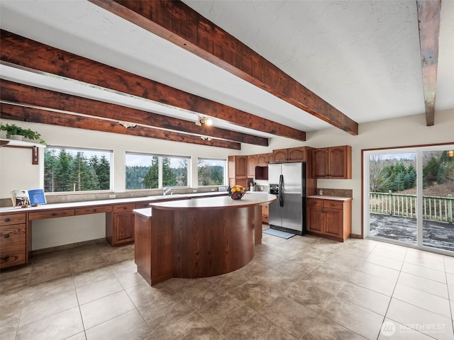 kitchen with beamed ceiling, brown cabinets, stainless steel refrigerator with ice dispenser, a center island, and light countertops