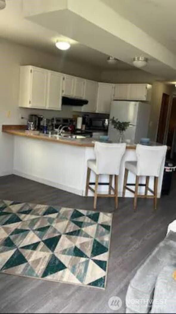 kitchen with a breakfast bar, freestanding refrigerator, dark wood-type flooring, under cabinet range hood, and white cabinetry